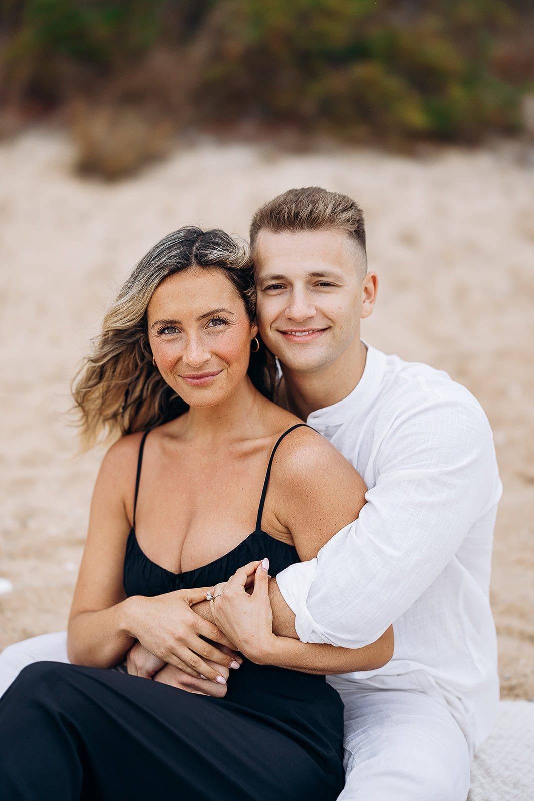 Couple seated on sandy beach, woman in black dress and man in white shirt embracing gently, natural expressions.