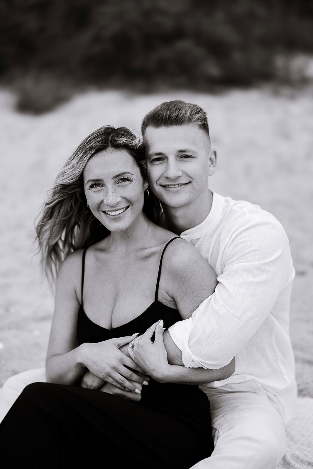 Black and white portrait of a couple sitting closely, the man embracing the woman from behind, both smiling gently, with a blurred natural background.