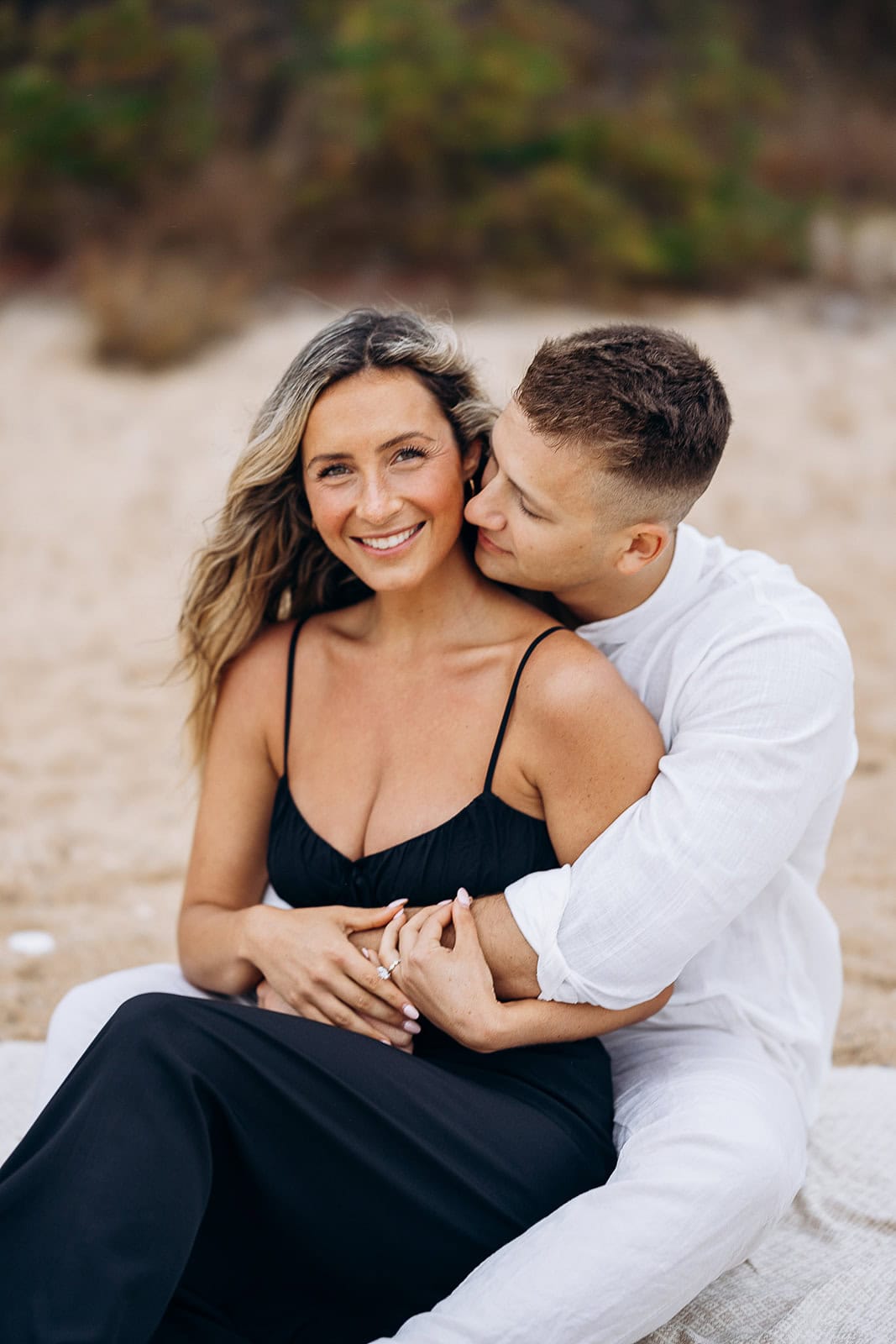 Couple seated on a sandy beach, the man in white shirt embraces the woman in a black dress, natural background blurred.