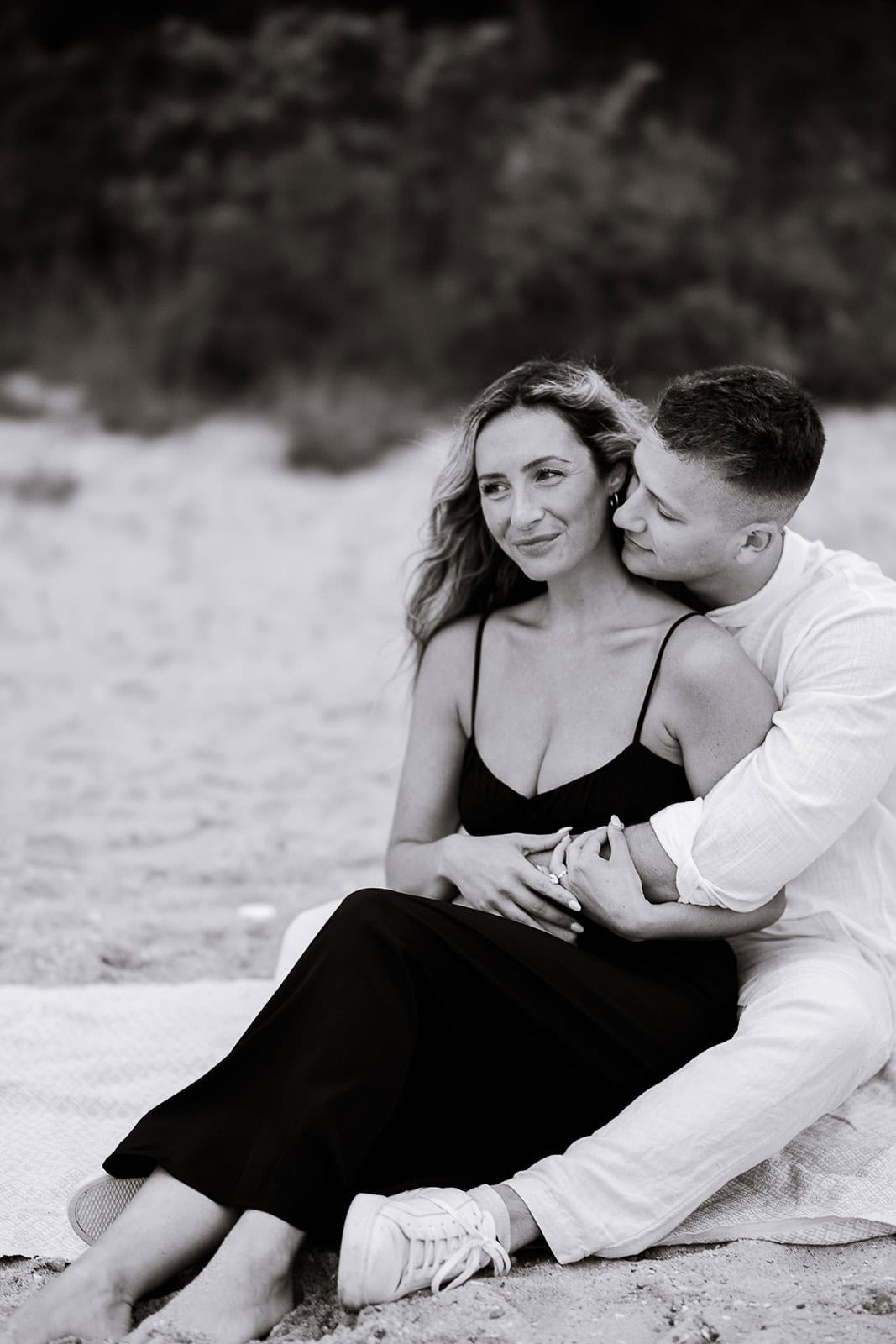 Black and white photo of a couple seated on a beach blanket, the man embraces the woman from behind as she looks away thoughtfully.