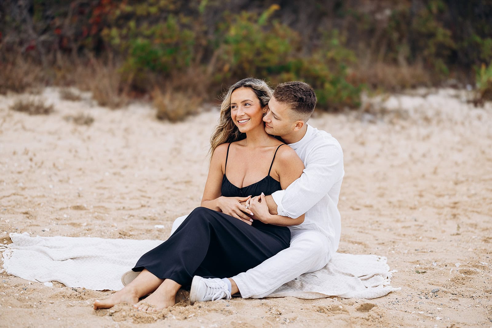 Couple seated on a blanket on sandy beach, man in white shirt and pants embracing woman in black dress, natural greenery in background.