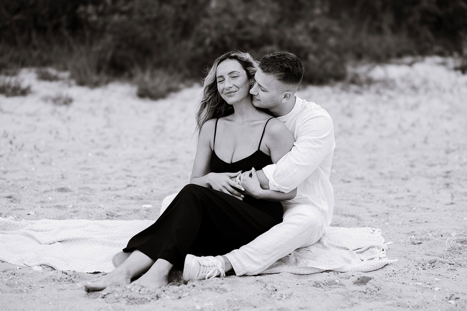 Black and white photo of a couple sitting on a blanket on the beach, the man embracing the woman from behind as she closes her eyes with a soft smile.