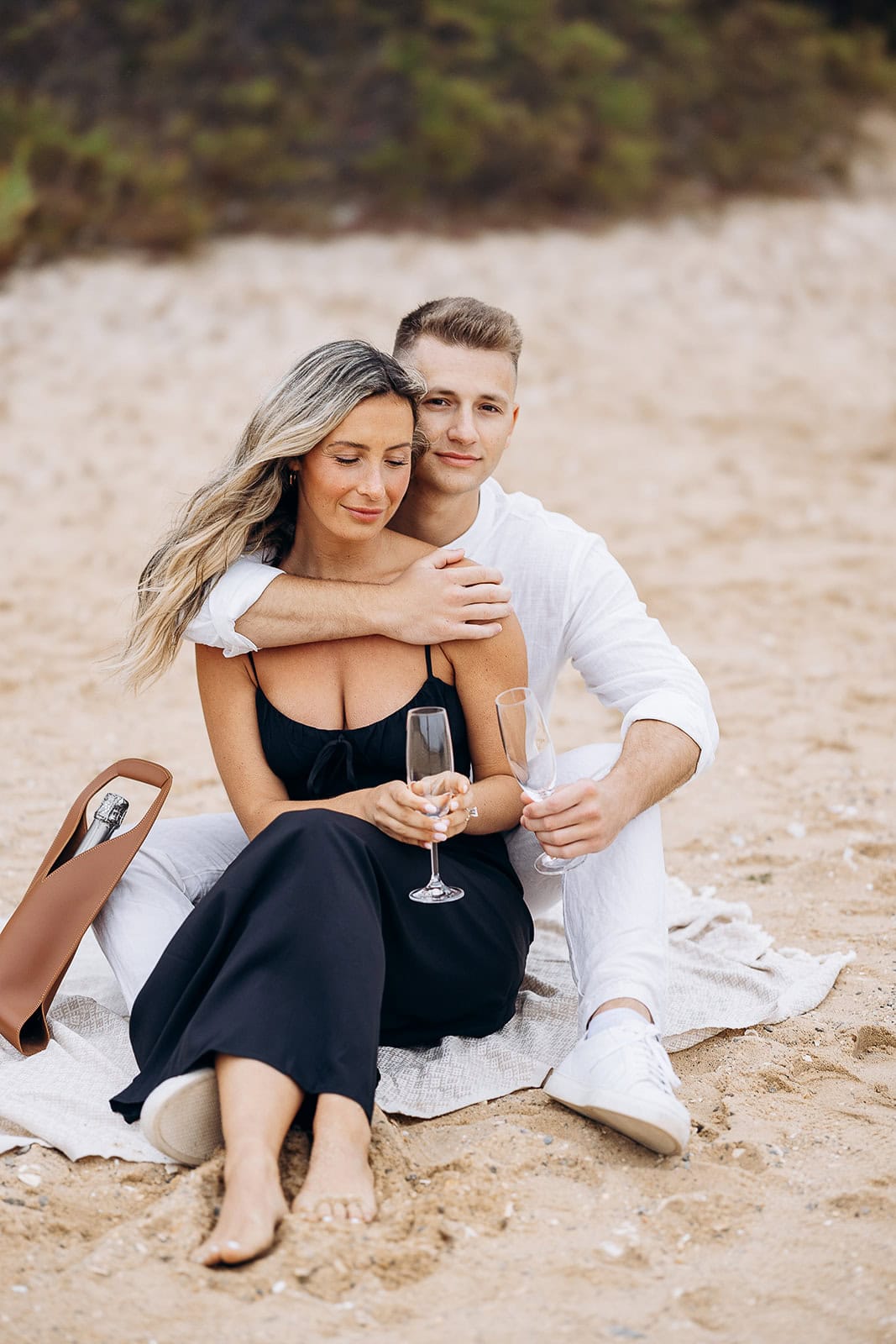 Couple seated on a sandy beach, the man in white embracing the woman in a black dress, holding champagne flutes with a bottle in a leather carrier nearby.