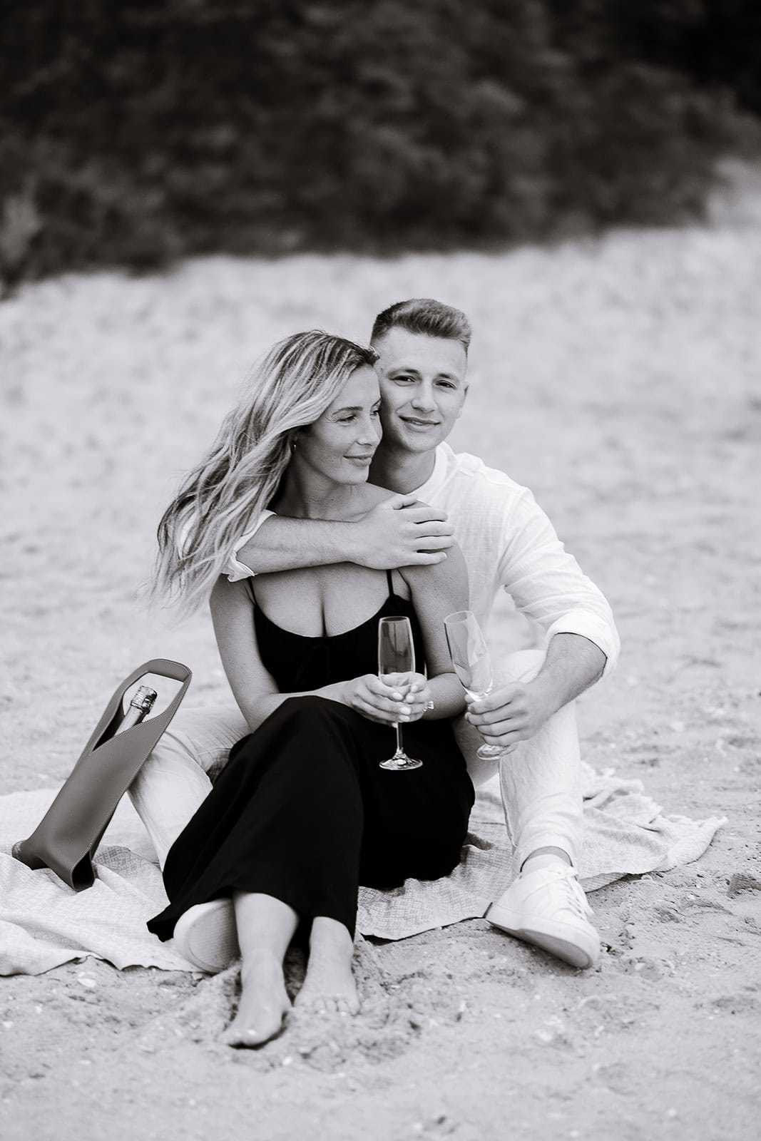 Black and white photo of a couple sitting on a beach blanket, the man in white embraces the woman in a black dress, both holding champagne flutes.