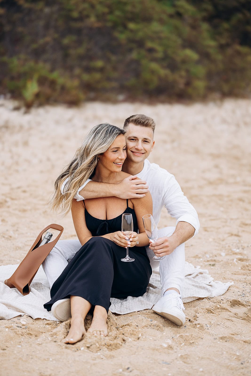 Couple seated on a beach blanket on sand, woman in black dress holding champagne glass, man in white outfit embracing her from behind with empty glass, champagne bottle in a tan leather carrier nearby.