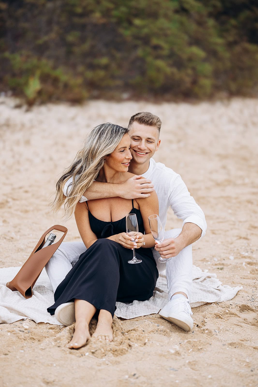 Couple seated on a sandy beach with a blanket, holding champagne flutes, a leather wine carrier with a bottle beside them.