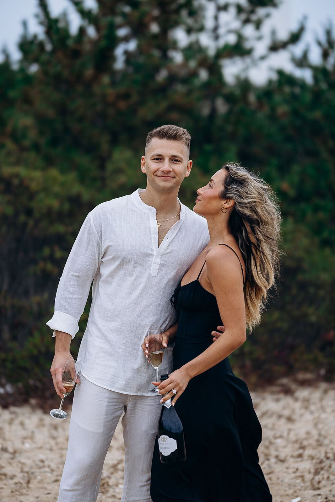 Couple standing on rocky ground with green foliage behind, man in white linen outfit holding champagne glass, woman in black dress holding champagne bottle and glass.