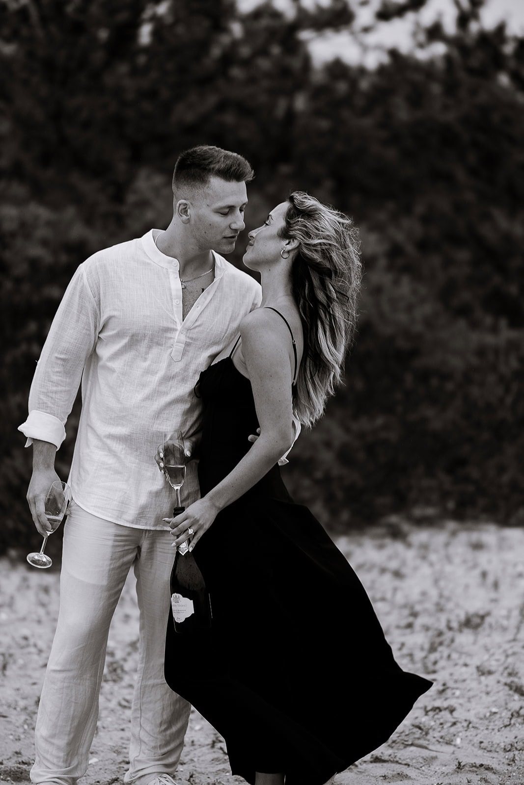 Couple standing close on sandy ground with trees blurred in the background, holding champagne glasses and bottle, poised for an intimate moment.