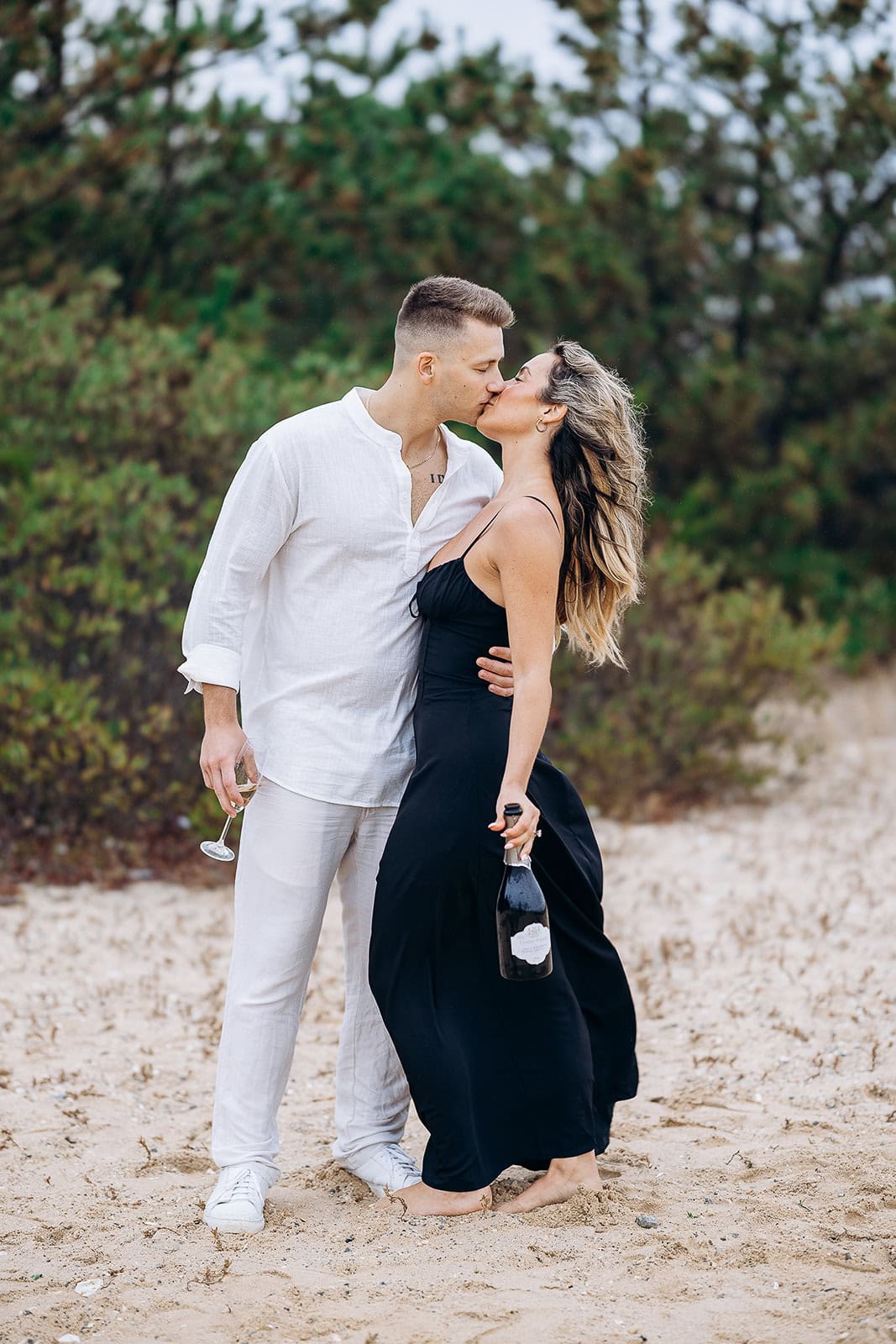 Couple kissing on sandy beach with greenery behind, woman in black dress holding champagne bottle, man in white linen outfit holding glass.