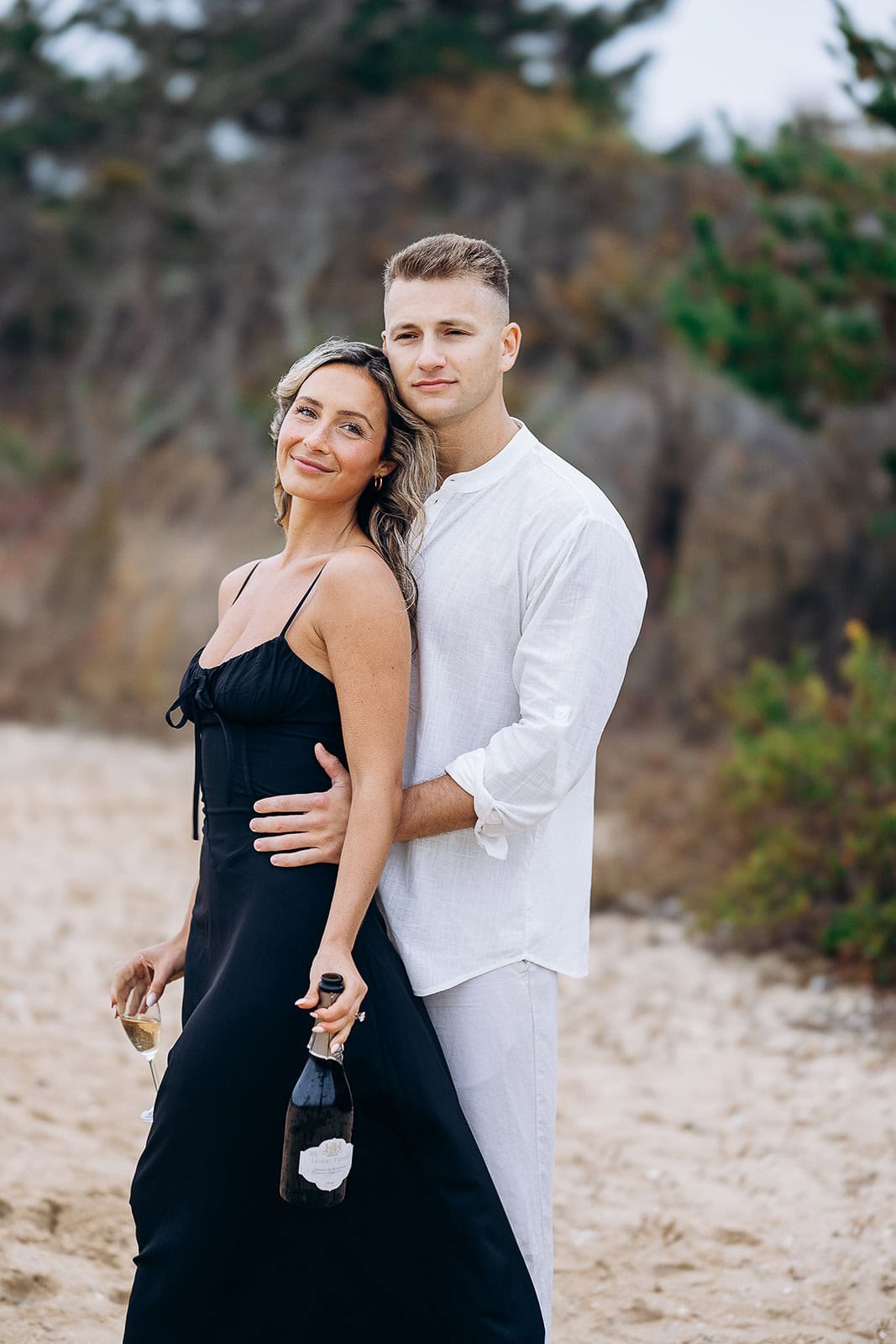Couple standing on a sandy beach with rocky and green foliage background; woman in a black dress holding a champagne bottle and glass, man in white shirt embracing her.