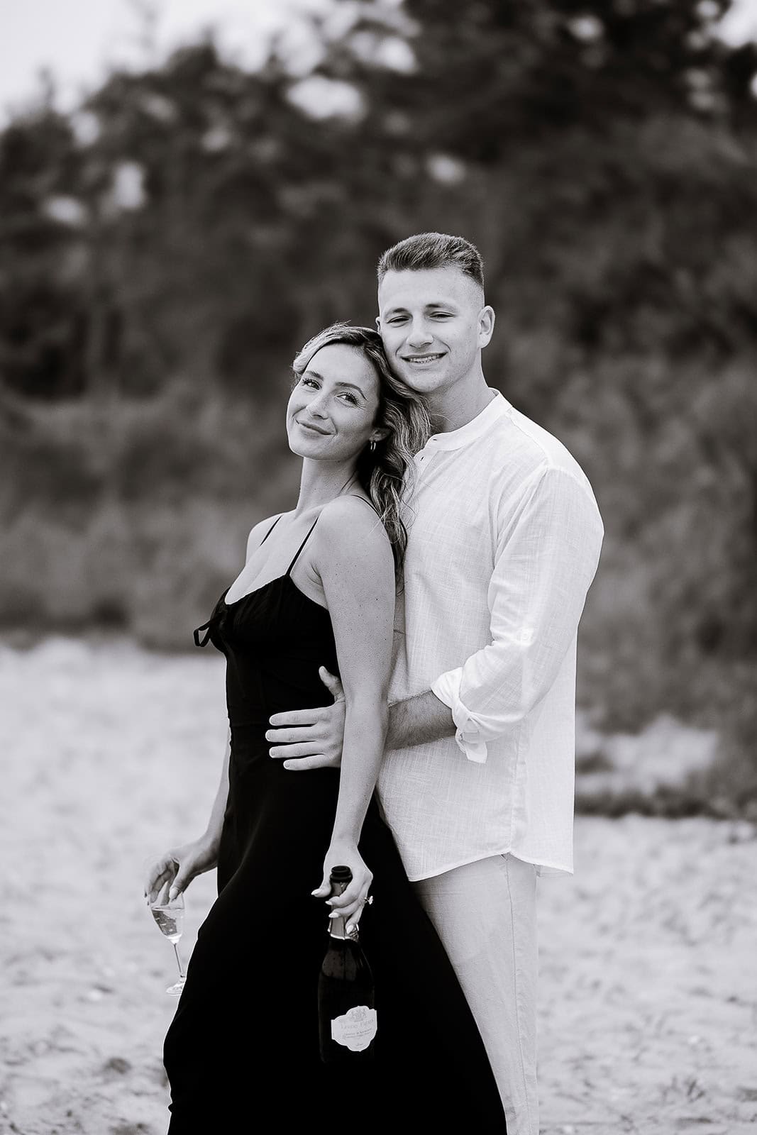 Black-and-white photo of a couple standing on a sandy beach, the man in a white shirt embracing the woman in a black dress holding a champagne bottle and glass.