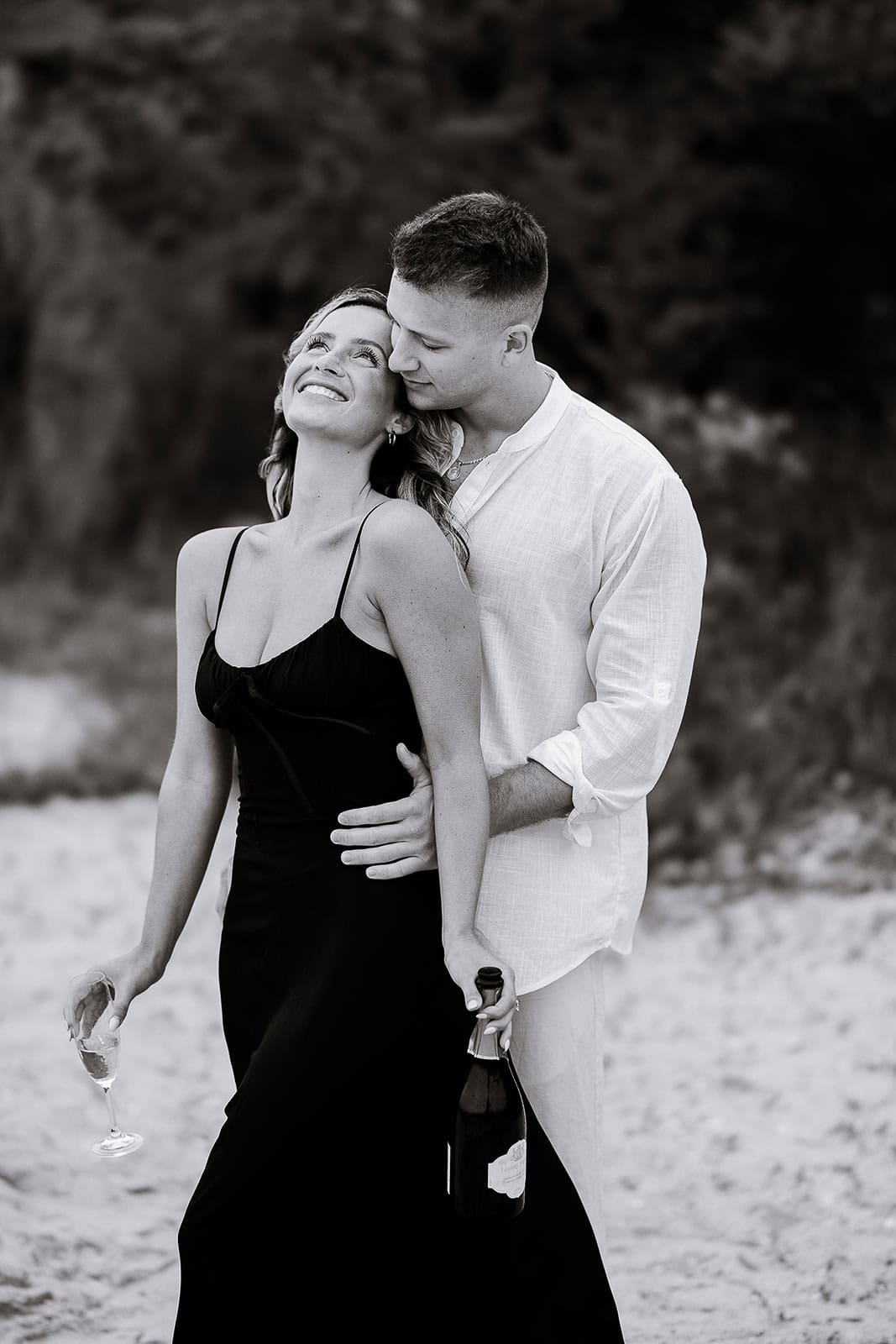 Black and white photo of a couple on a sandy beach; woman in black dress holding a champagne glass, man in white shirt embracing her while holding a champagne bottle.
