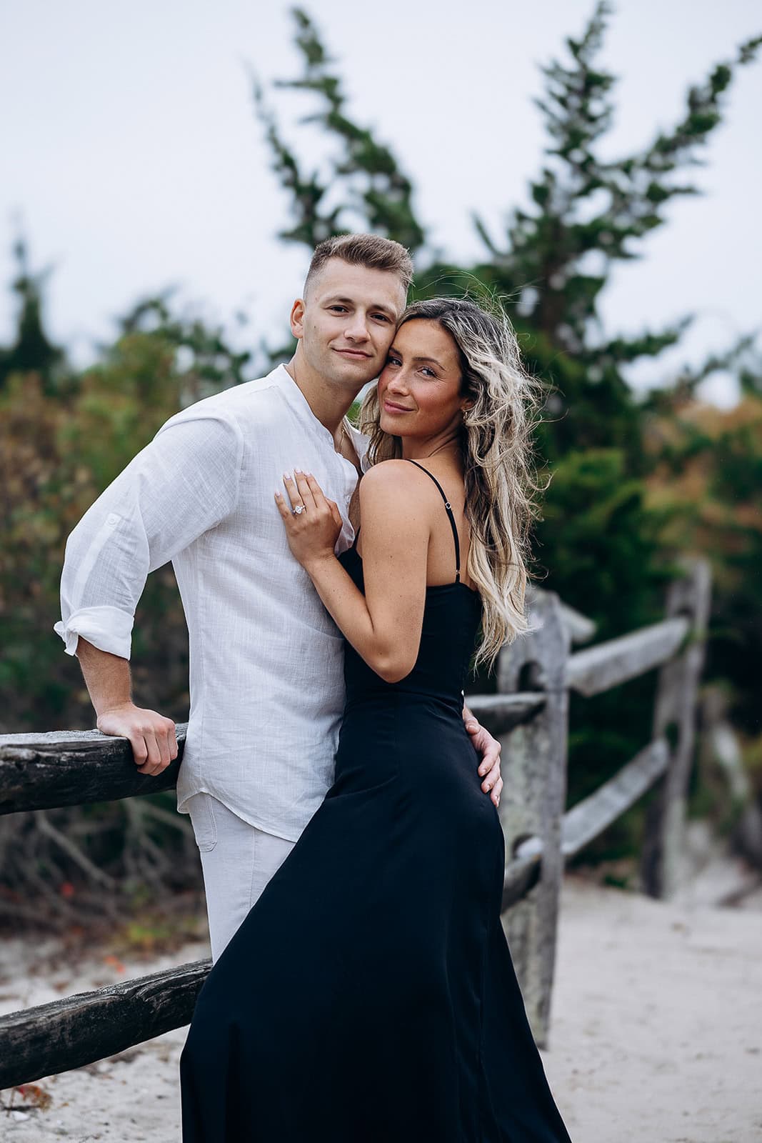 Couple embracing near a weathered wooden fence with greenery in the background at Barnegat Light, NJ.