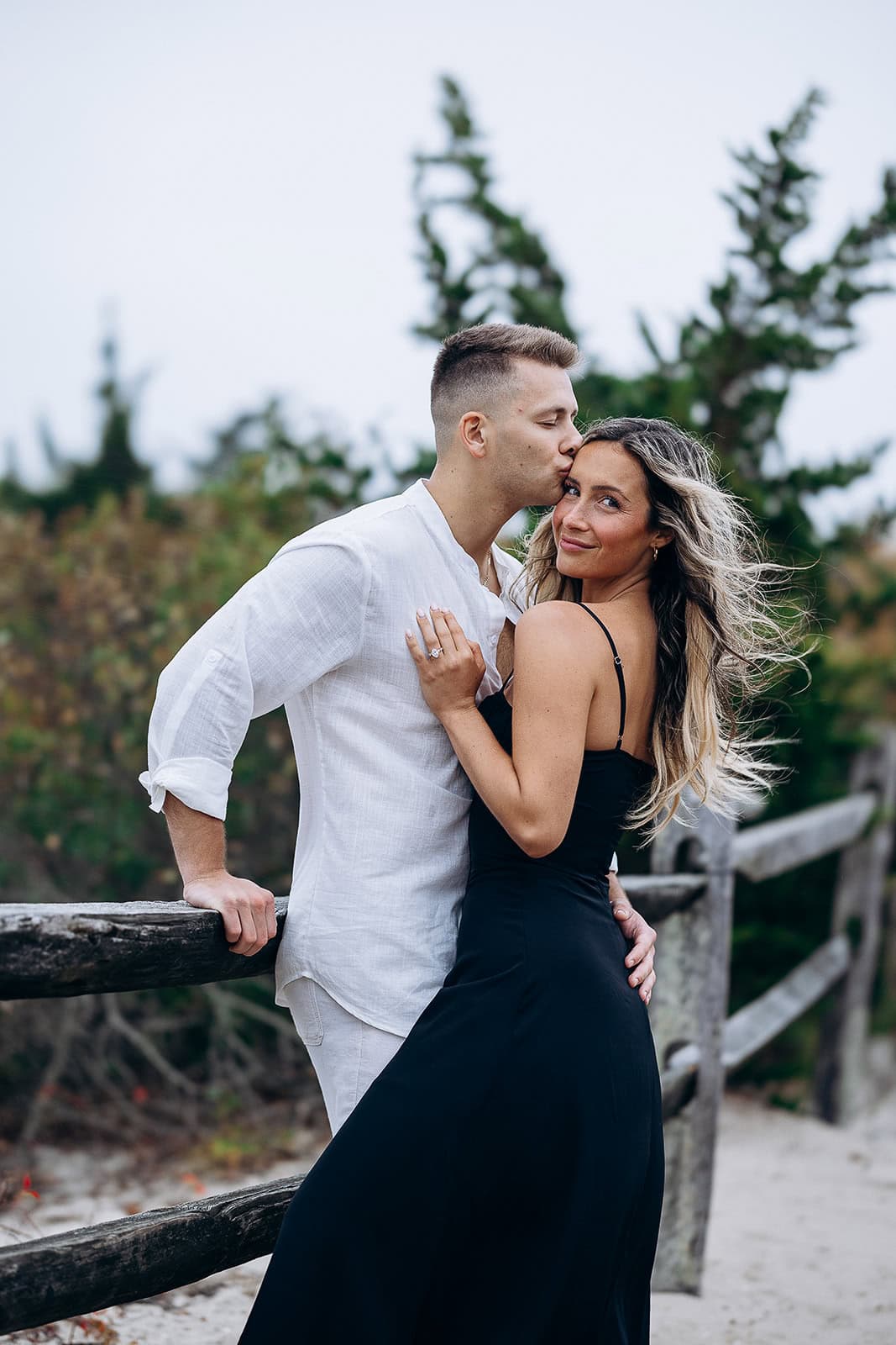 Couple embracing by a weathered wooden fence on a sandy path, man in white shirt kissing woman in black dress on forehead, greenery in background.