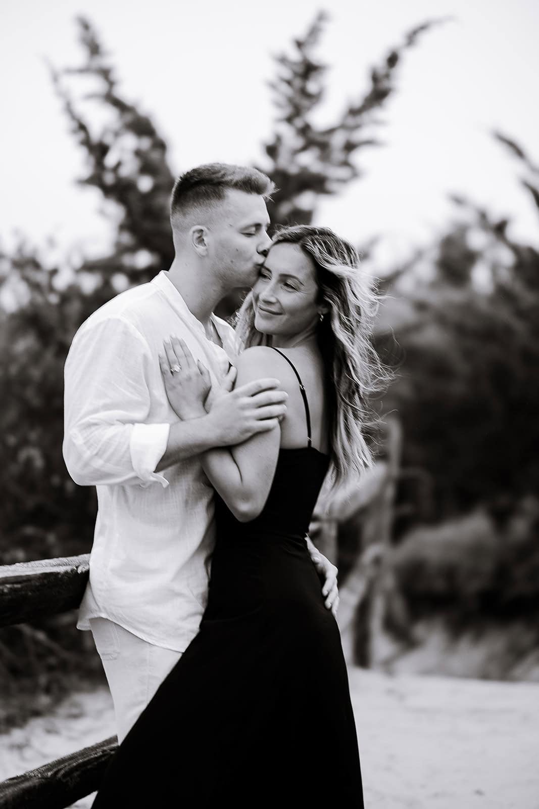 Black and white image of a man in a white shirt gently kissing a woman in a black dress on the forehead near a wooden fence with blurred trees in the background.