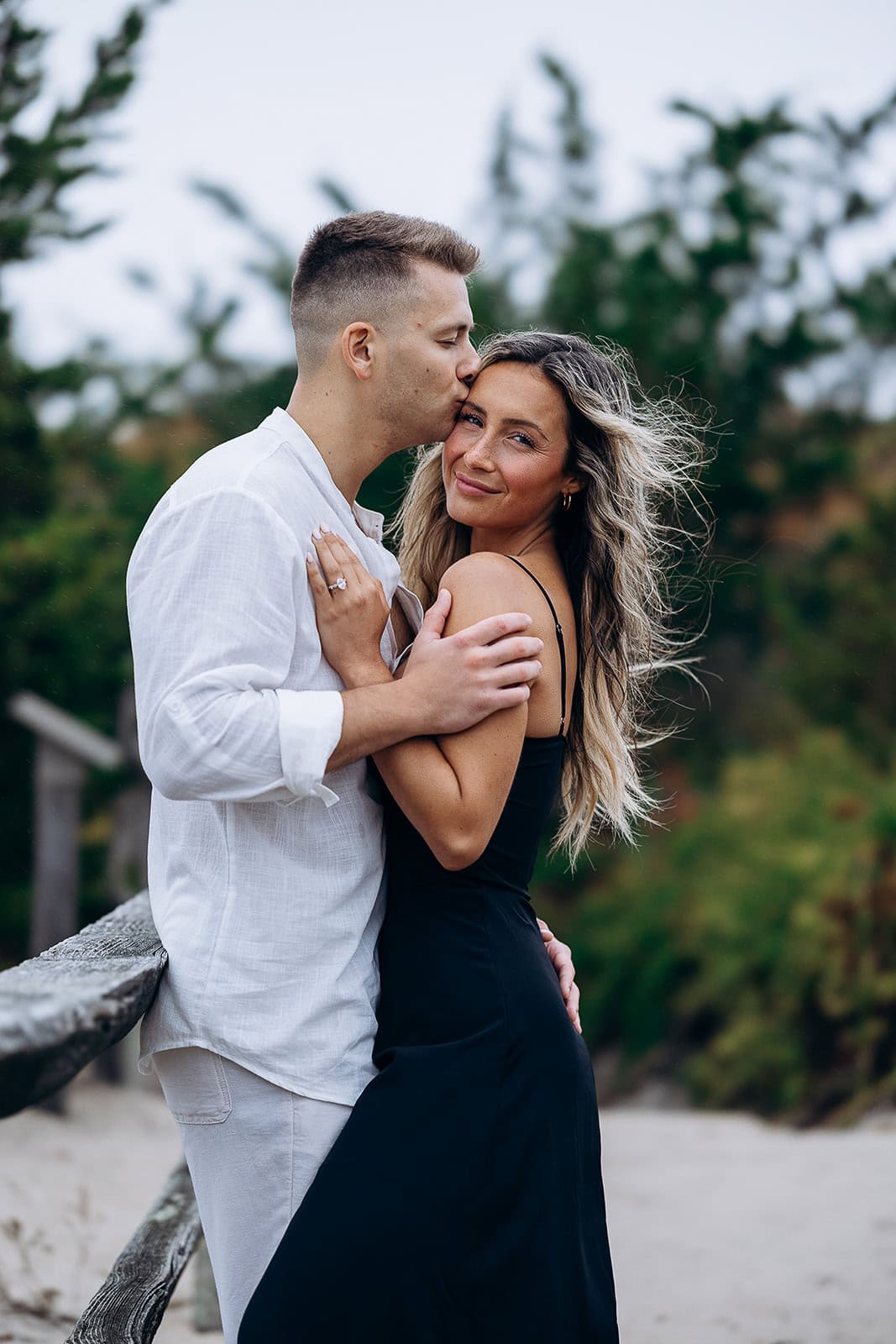 Man in white shirt kissing woman in black dress on forehead by wooden railing with blurred green foliage background.