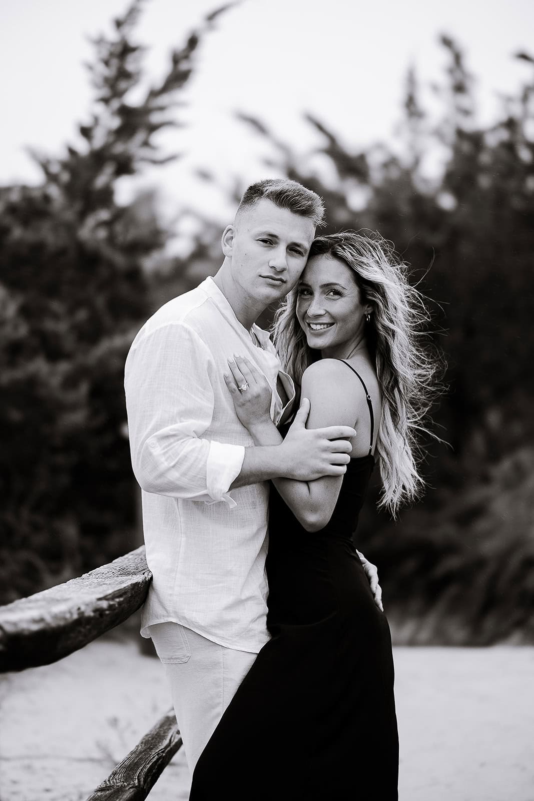 Black and white photo of a couple embracing near a wooden fence with blurred trees in the background, showcasing natural intimacy and texture.