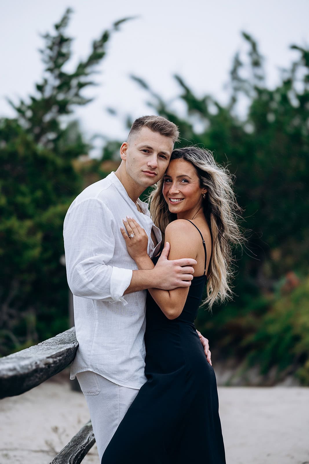 Couple embracing near rustic wooden fence with green trees in background, outdoors in natural setting.