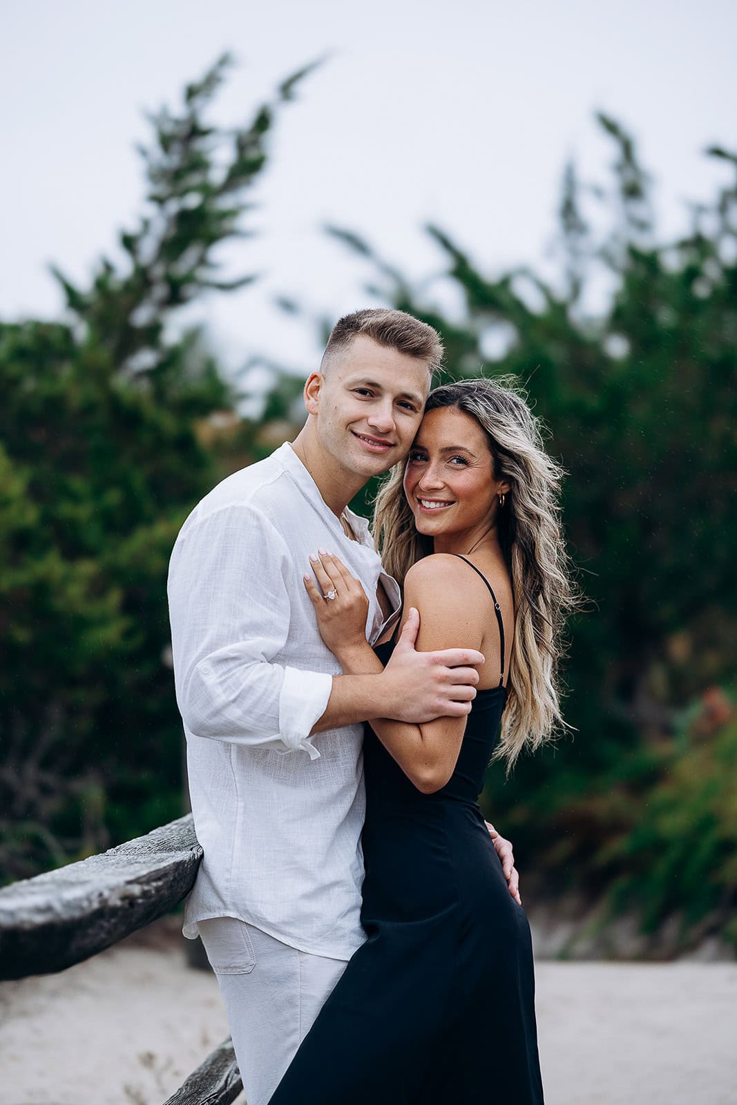 Couple embracing near wooden fence with blurred green foliage background, showcasing natural light and intimate pose.