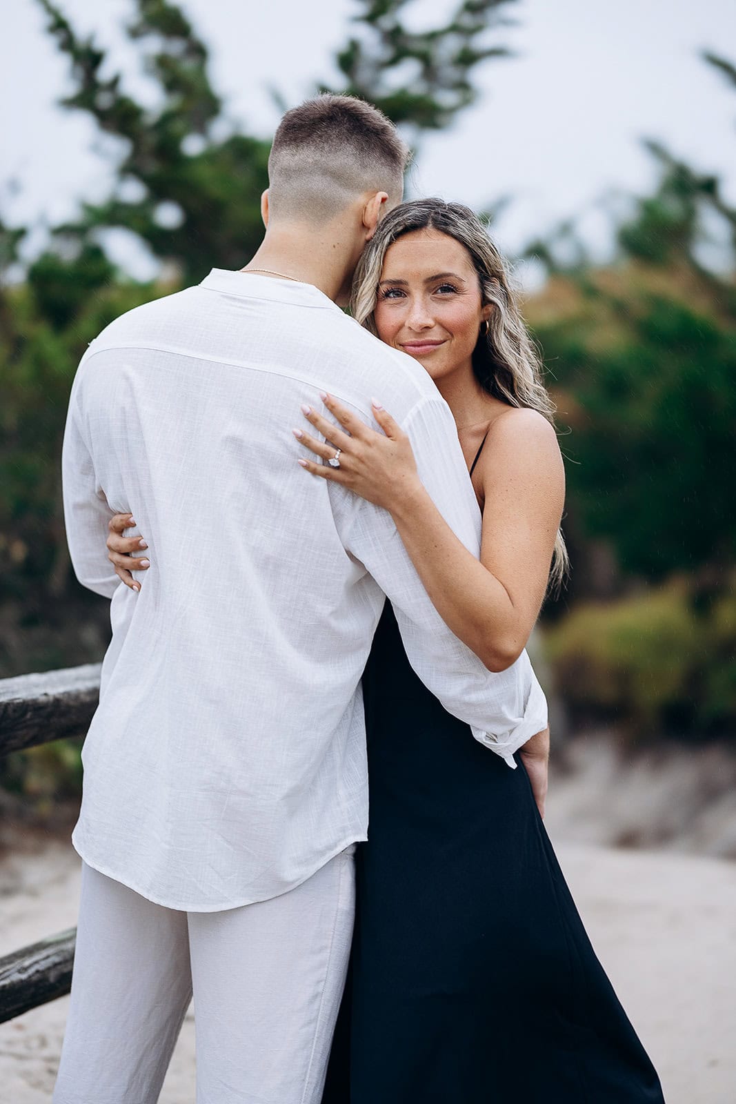 Couple embracing outdoors with woman facing camera, man in white shirt with back to camera, natural greenery background.