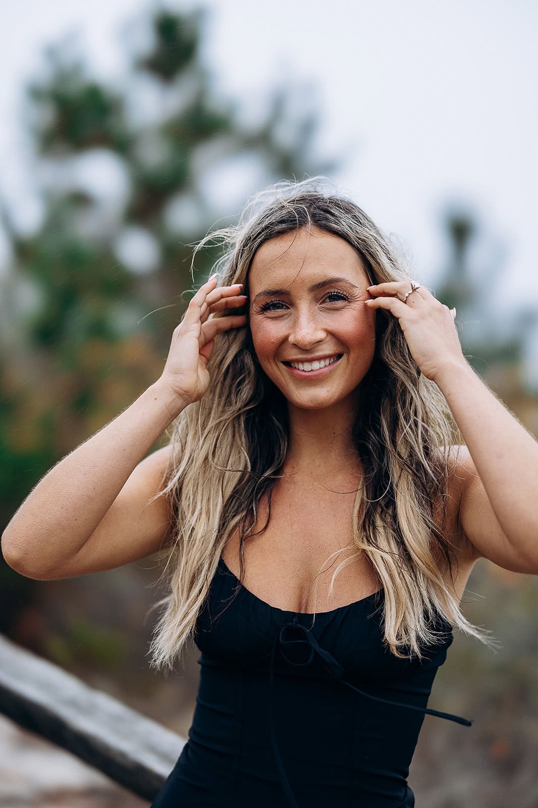 Close-up of a smiling woman with long blonde hair, wearing a black sleeveless top, outdoors with blurred trees in the background.