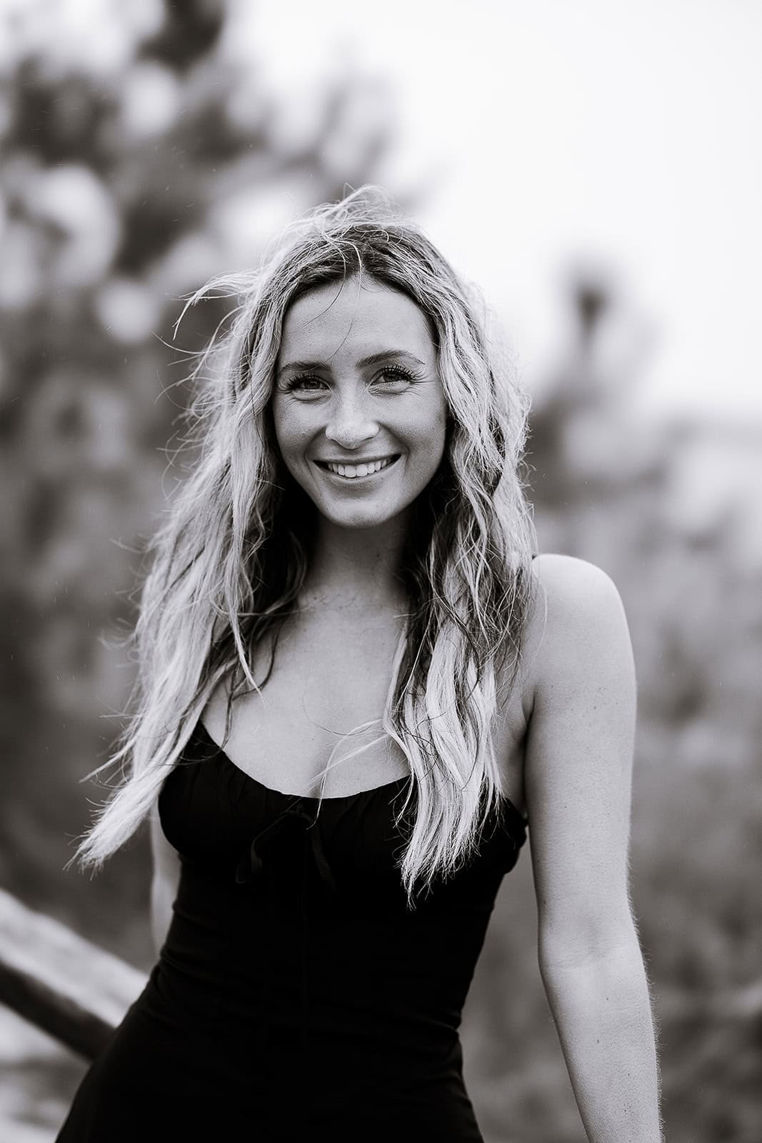 Black and white close-up portrait of a woman with long wavy hair smiling, blurred natural background.