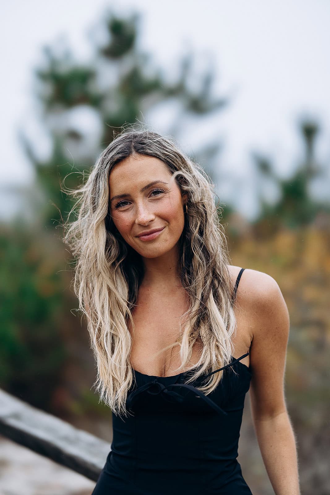 Close-up portrait of a woman with long wavy blonde hair wearing a black dress, standing outdoors with blurred trees in the background.
