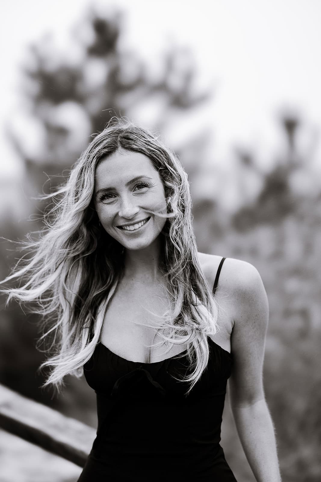 Black-and-white portrait of a woman with long hair flowing in the wind, smiling gently, wearing a sleeveless black top with blurred natural background.