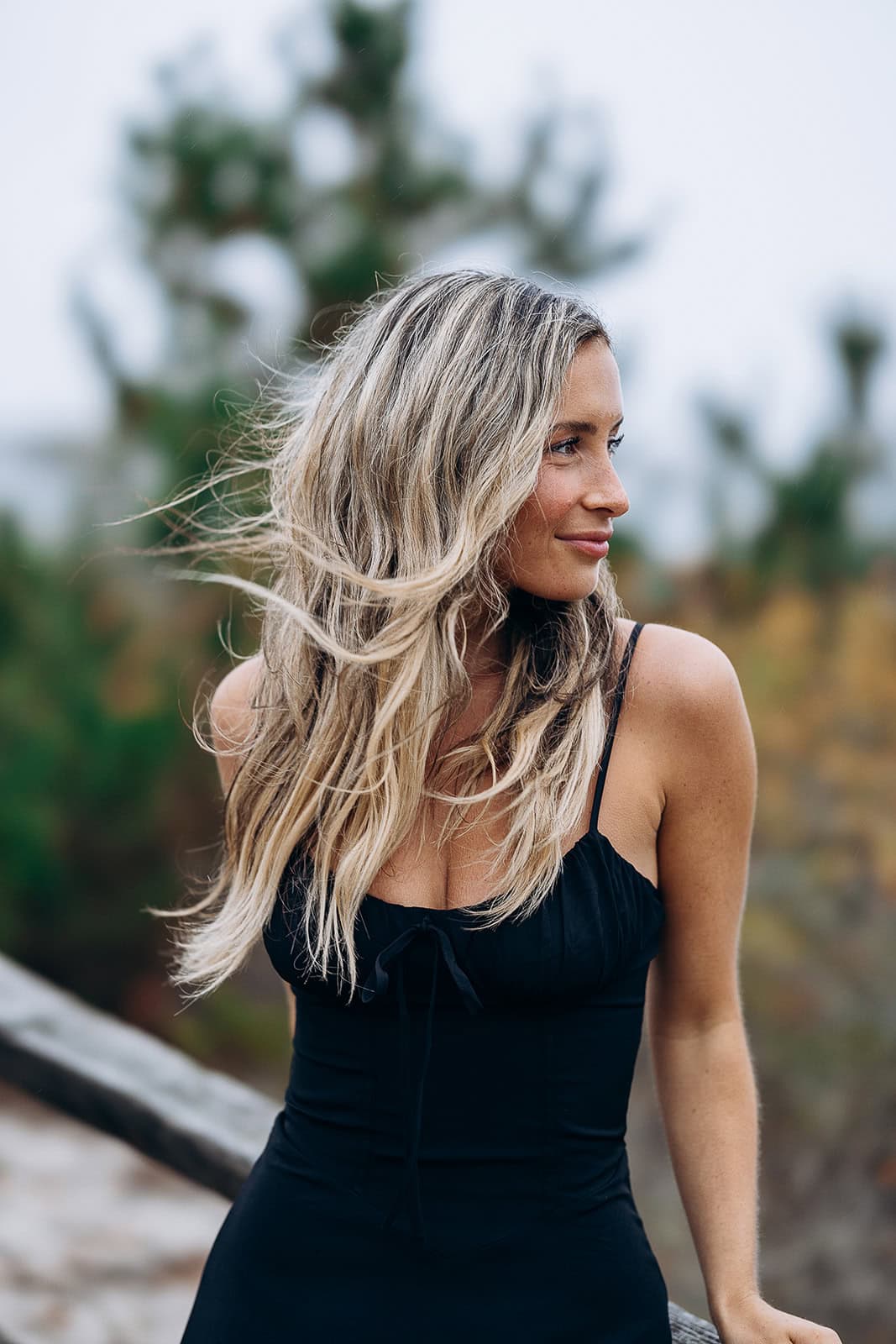 Woman with long blonde hair in a black dress standing outdoors near wooden railing with blurred greenery in background at Barnegat Light, NJ.