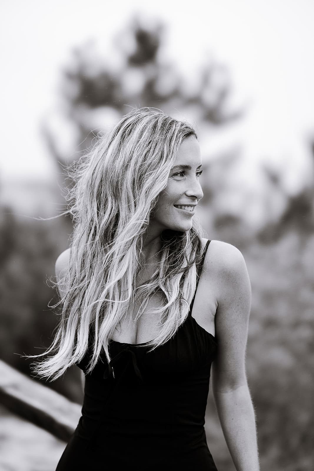 Black and white portrait of a woman with long flowing hair looking to the side, wearing a sleeveless black dress, with blurred natural background.
