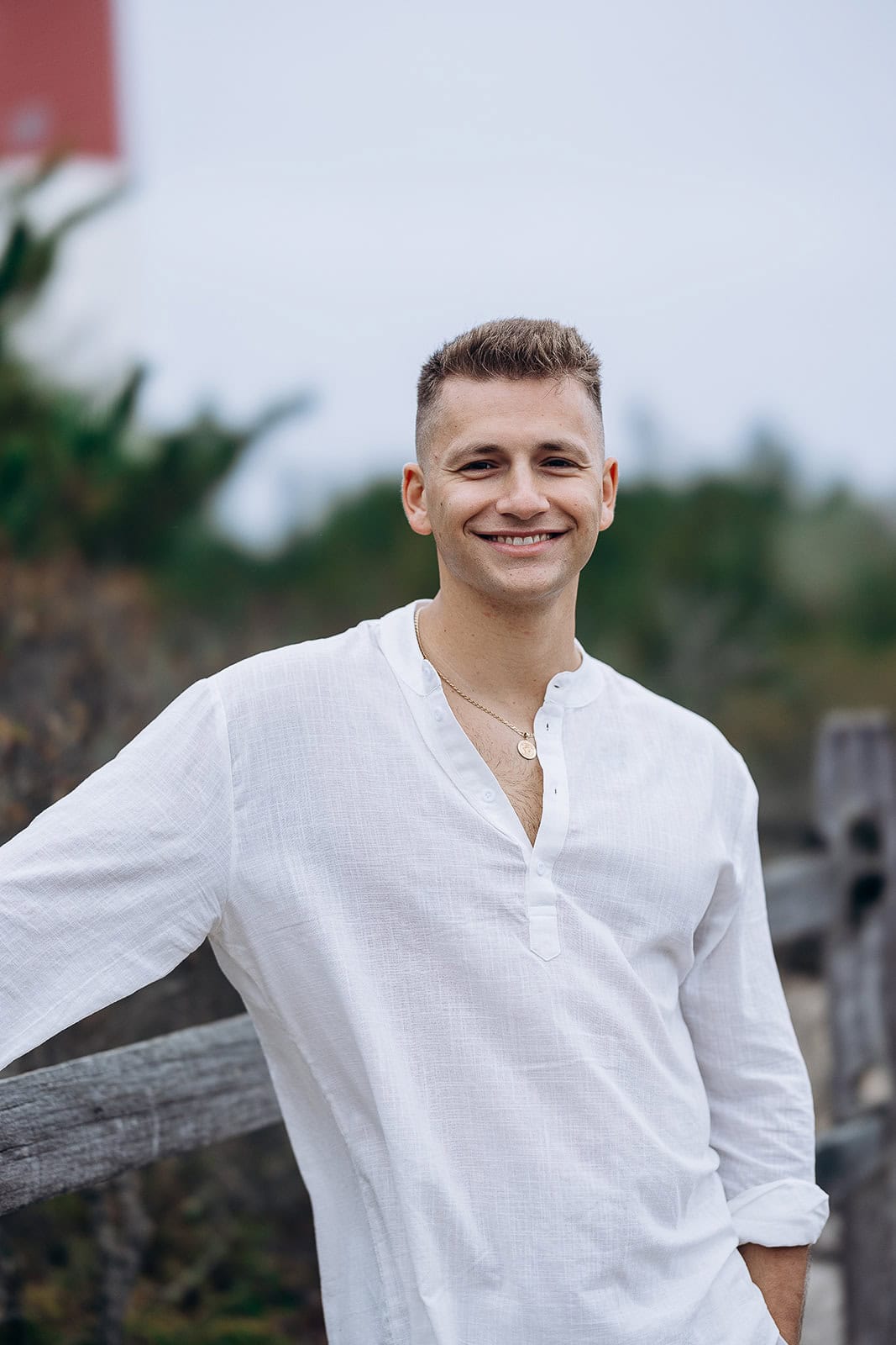 Smiling man in a white linen shirt leaning on a rustic wooden fence with blurred greenery in the background.
