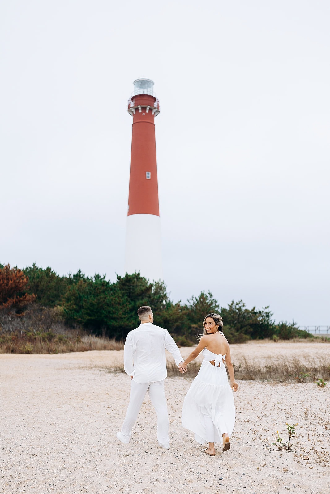 Couple dressed in white holding hands walking on sandy beach with Barnegat Light Lighthouse and greenery in the background, Barnegat Light, NJ.