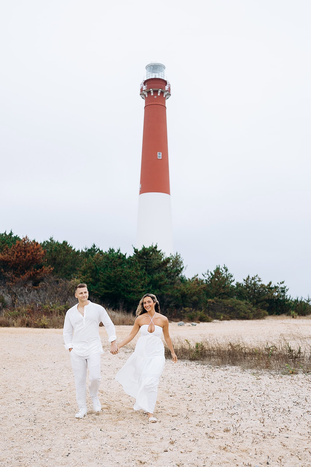 Couple holding hands walking on sandy beach near Barnegat Light Lighthouse with greenery in background on overcast day