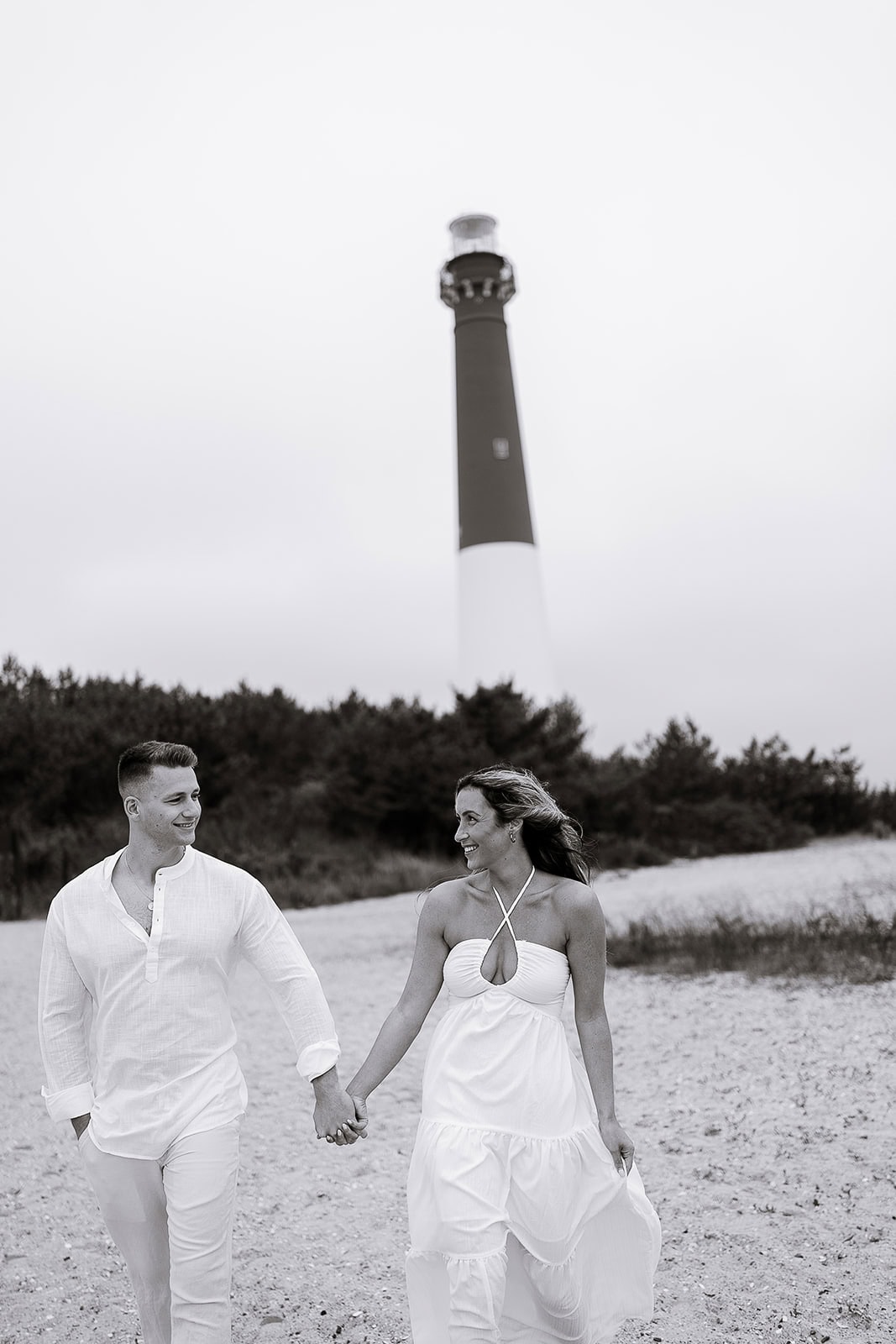 Black-and-white photo of a couple holding hands and walking on a beach with Barnegat Light Lighthouse in the background.