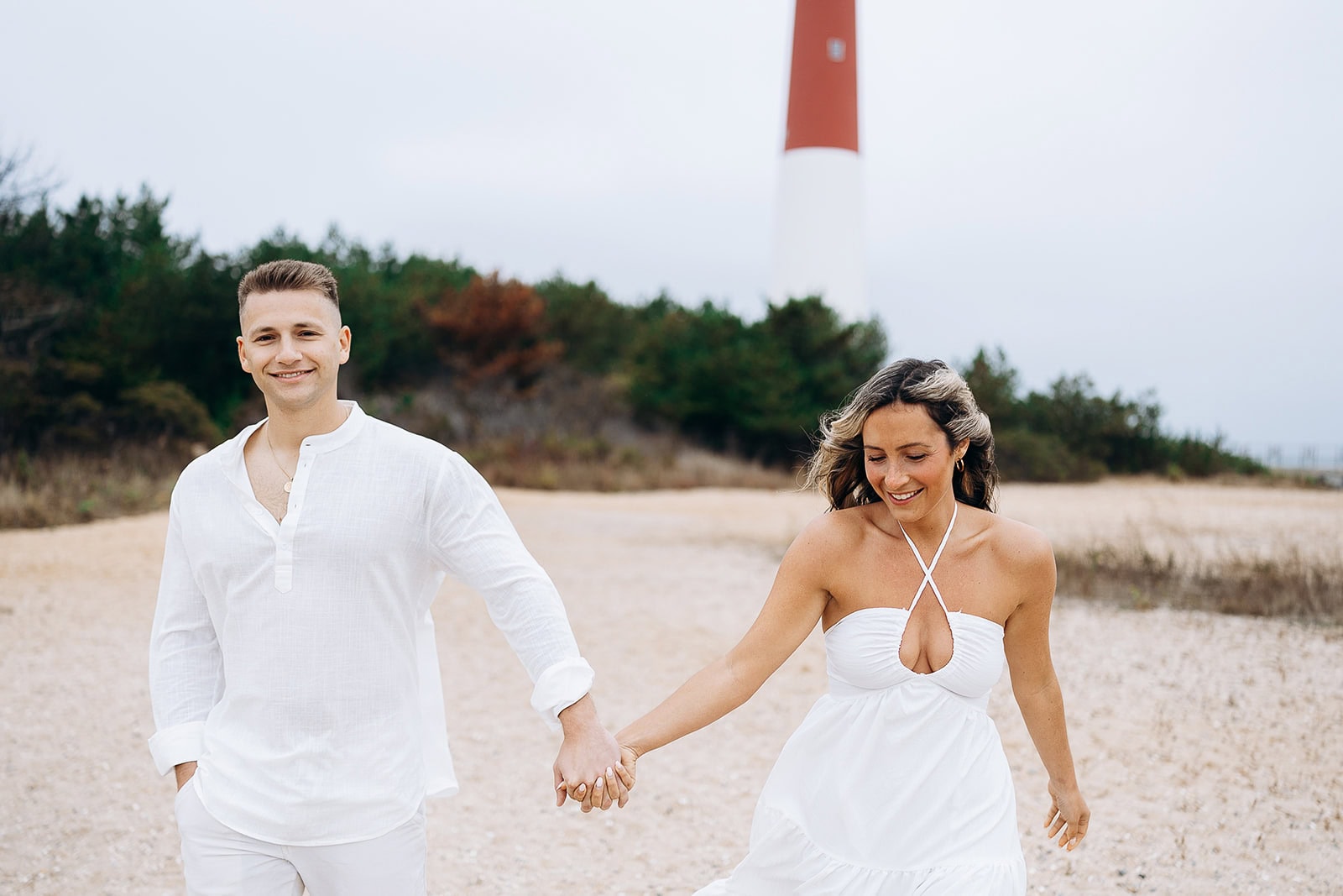 Couple dressed in white holding hands on a sandy beach with Barnegat Light lighthouse and greenery in the background.
