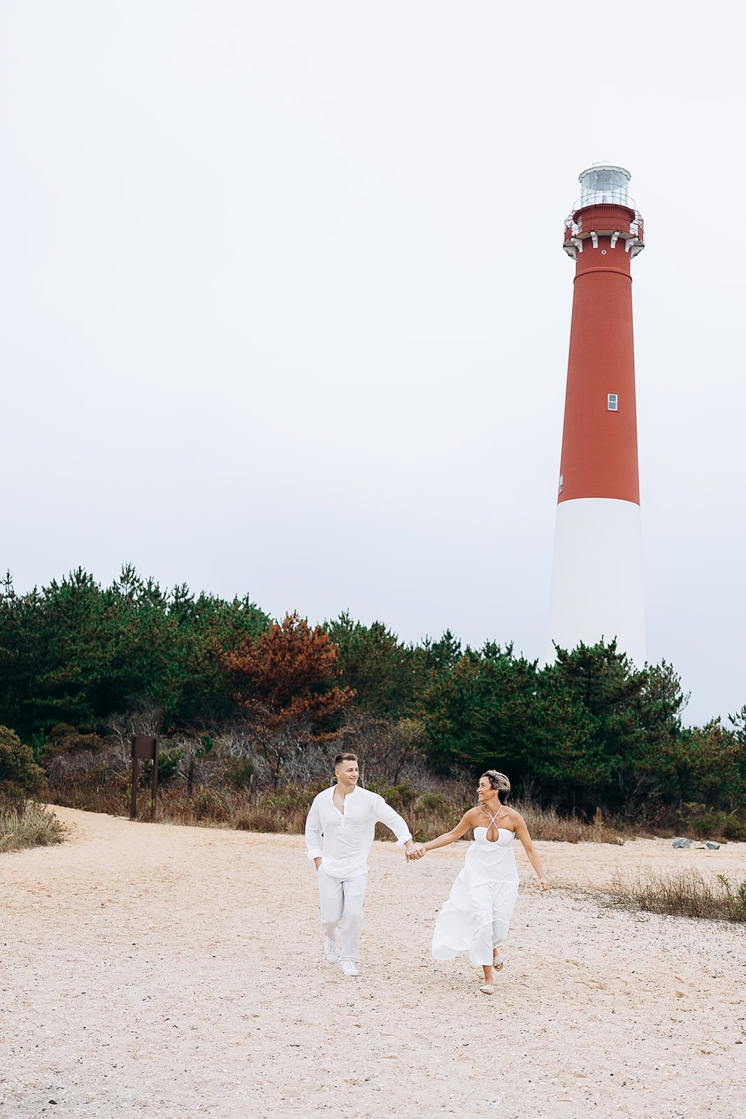 Couple dressed in white holding hands and walking on sandy beach with Barnegat Light lighthouse and green trees in the background.