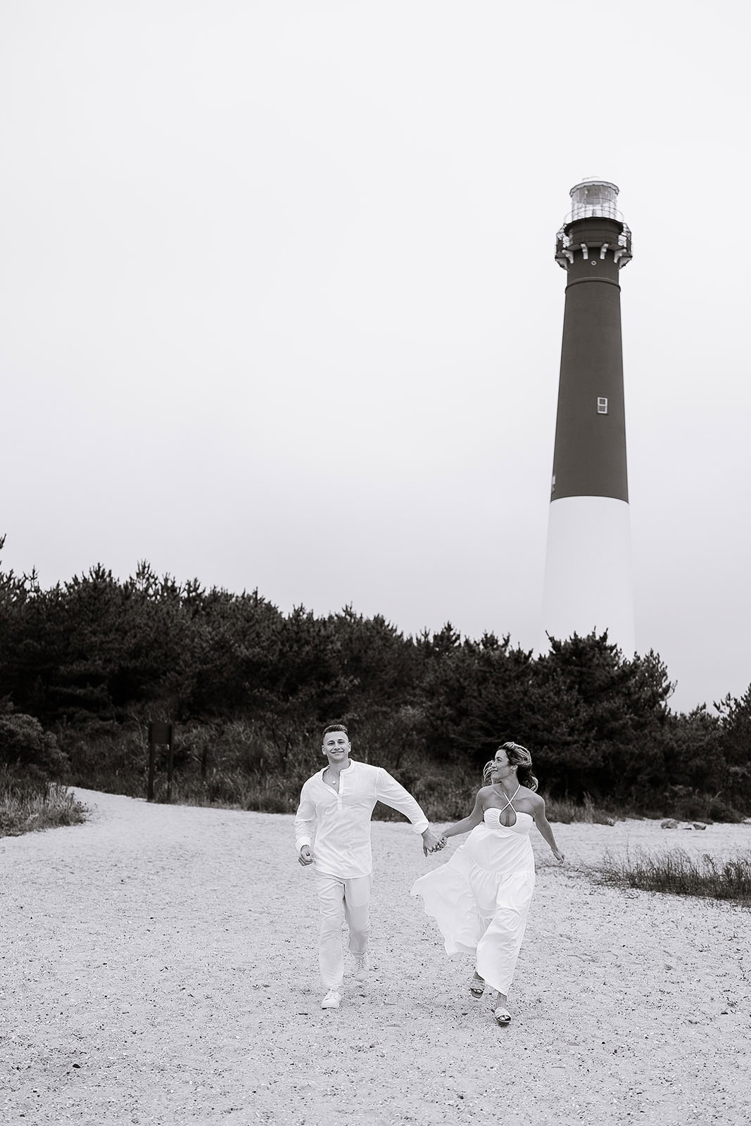 Couple holding hands and running on sandy path with Barnegat Light lighthouse and dense trees in the background, Barnegat Light, NJ.