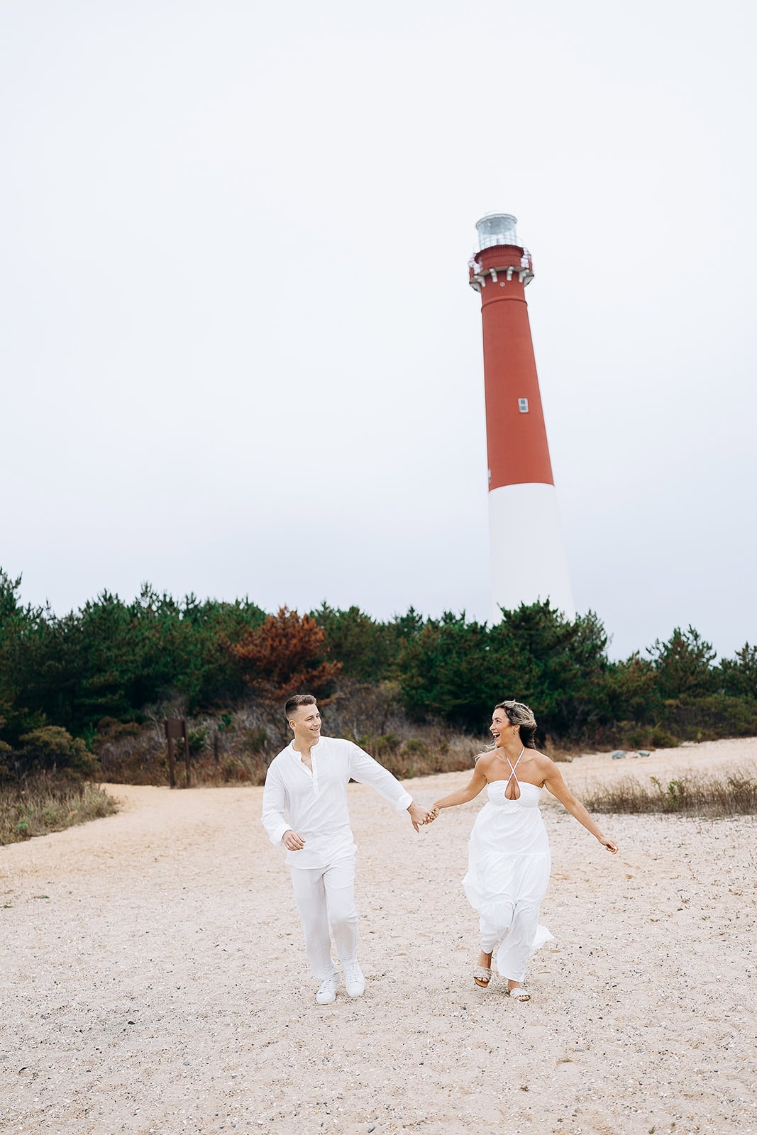 Couple in white attire holding hands and walking on sandy beach with Barnegat Light Lighthouse and greenery in the background.