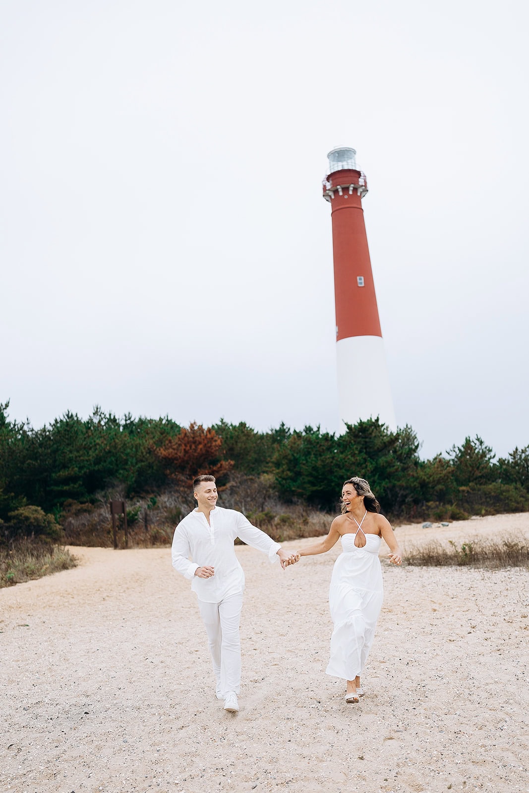 Couple in white clothing holding hands and running on sandy path with Barnegat Light Lighthouse and green trees in the background