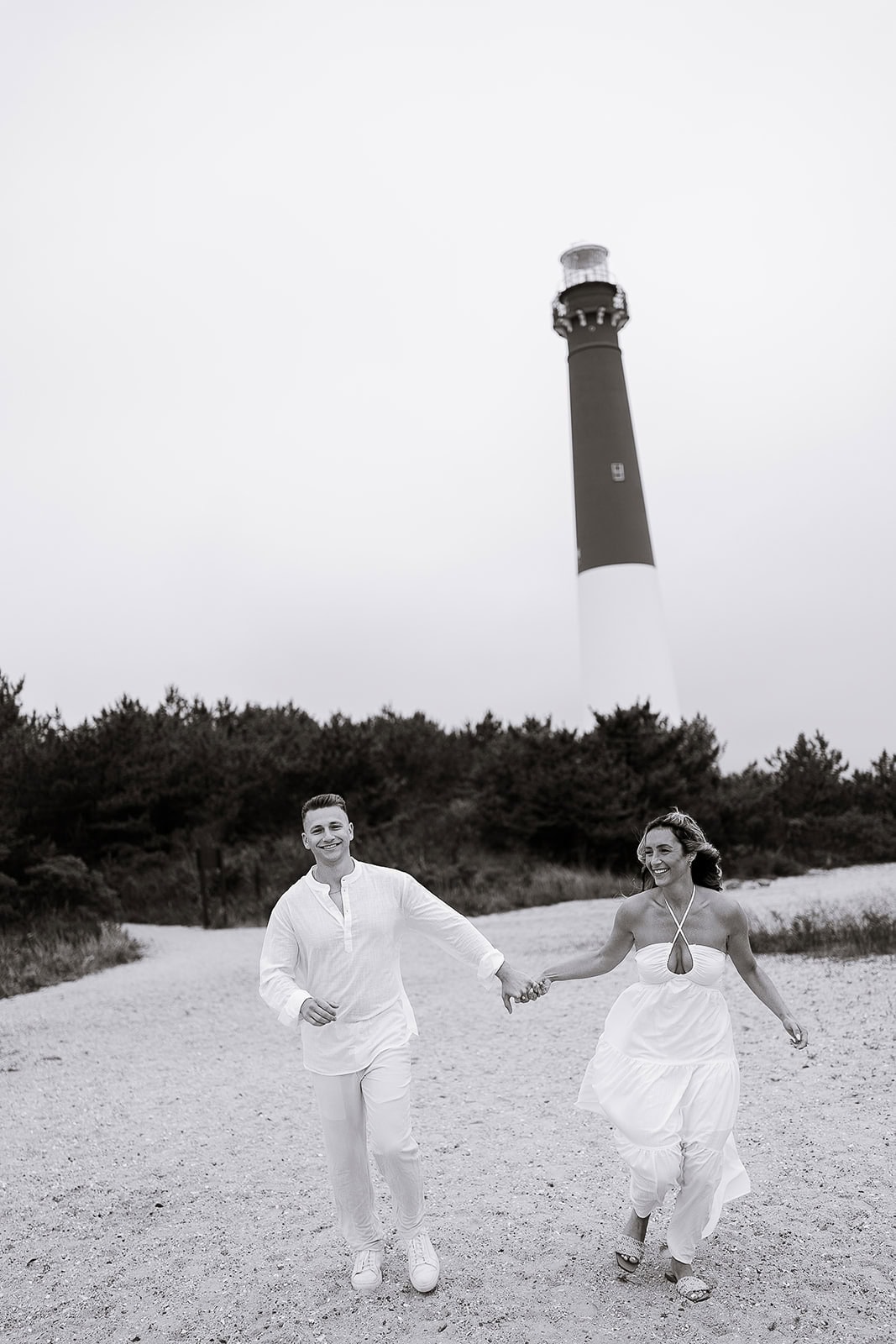 Couple dressed in white walking hand-in-hand on sandy path with Barnegat Light lighthouse and trees in background, Barnegat Light, NJ.
