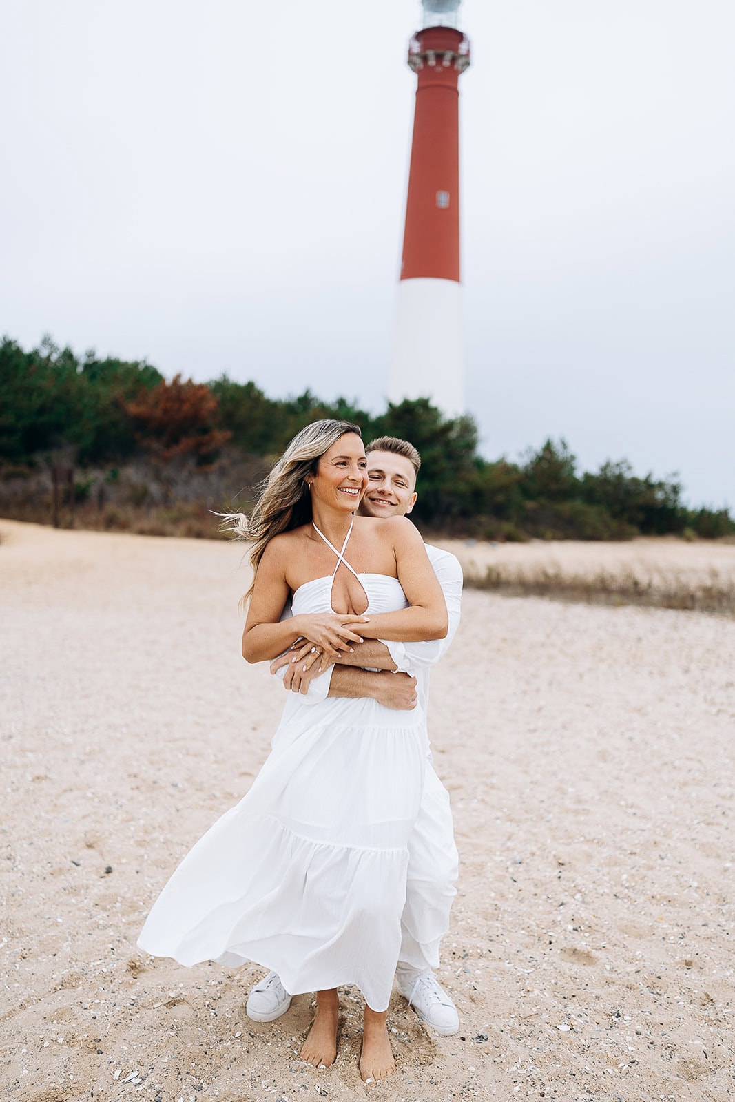 Couple in white clothing embracing on sandy beach with Barnegat Light lighthouse and greenery in the background