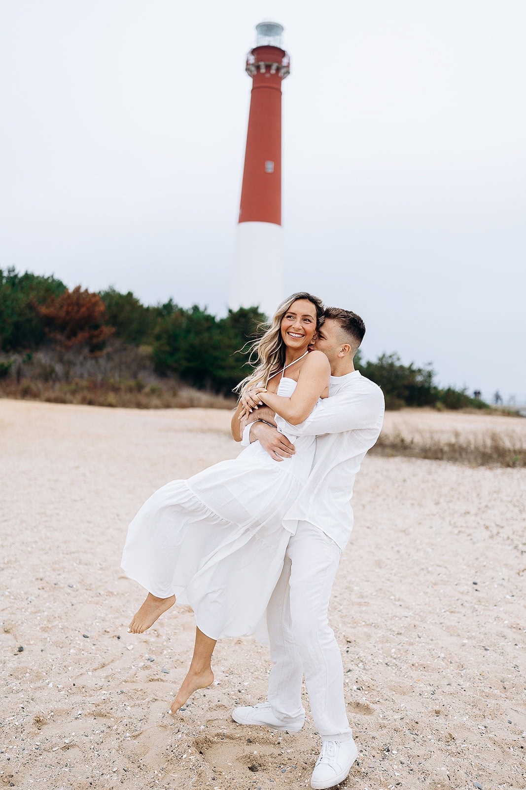 Couple in white clothing embracing on sandy beach with Barnegat Light Lighthouse in the background under overcast sky.