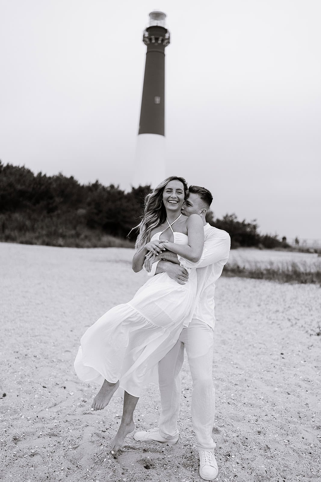 Couple in white attire embracing on sandy beach with Barnegat Light lighthouse in the background, captured in black and white.