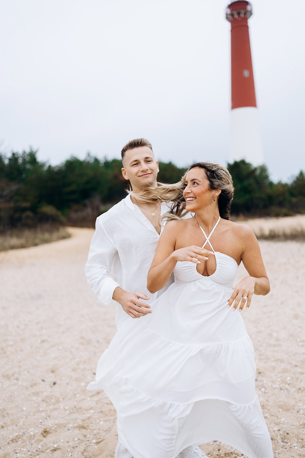 Couple dressed in white walking on sandy path near Barnegat Light Lighthouse with trees in the background.