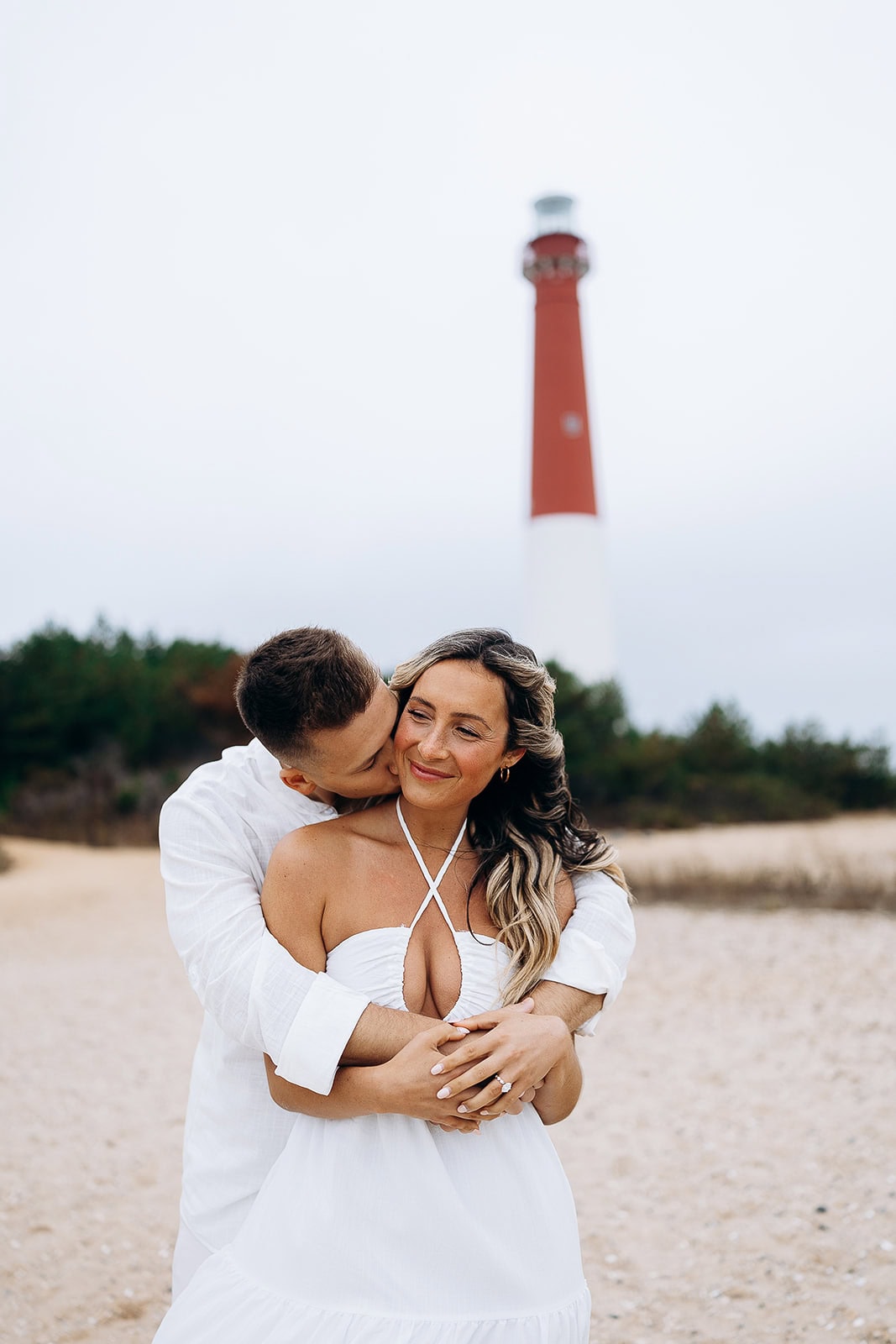 Couple in white clothing embracing on sandy beach with Barnegat Light lighthouse blurred in background