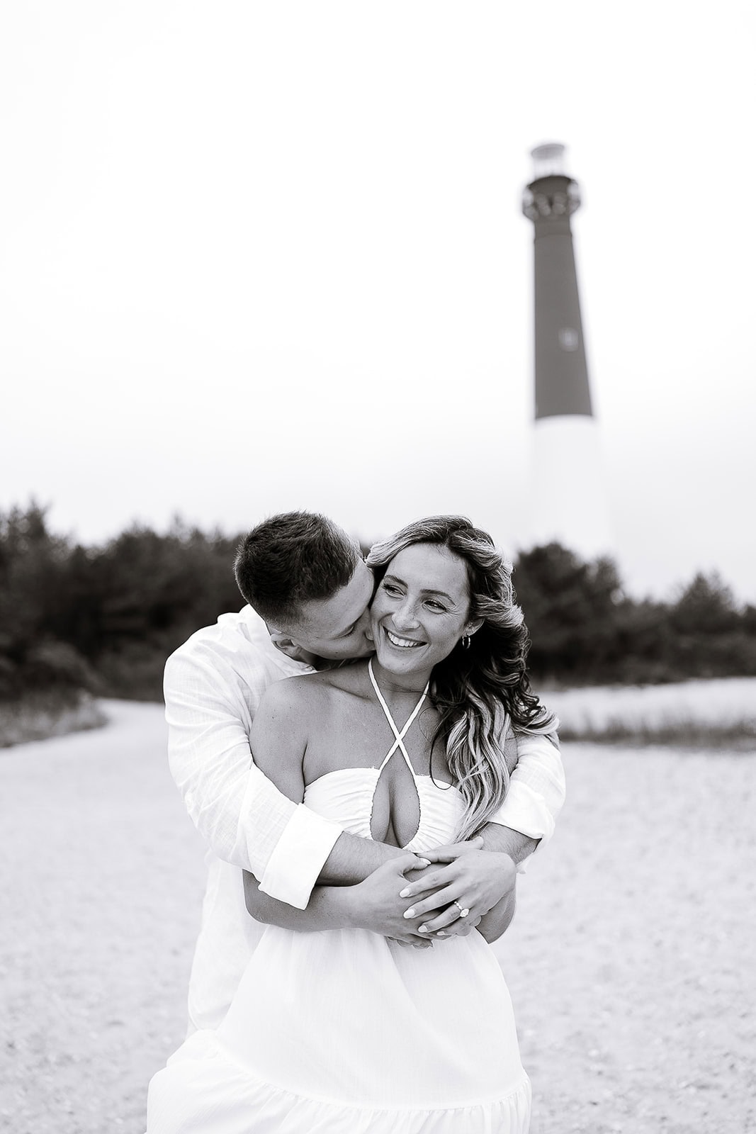 Couple in white attire sharing an intimate embrace on a sandy path with Barnegat Light lighthouse blurred in the background.