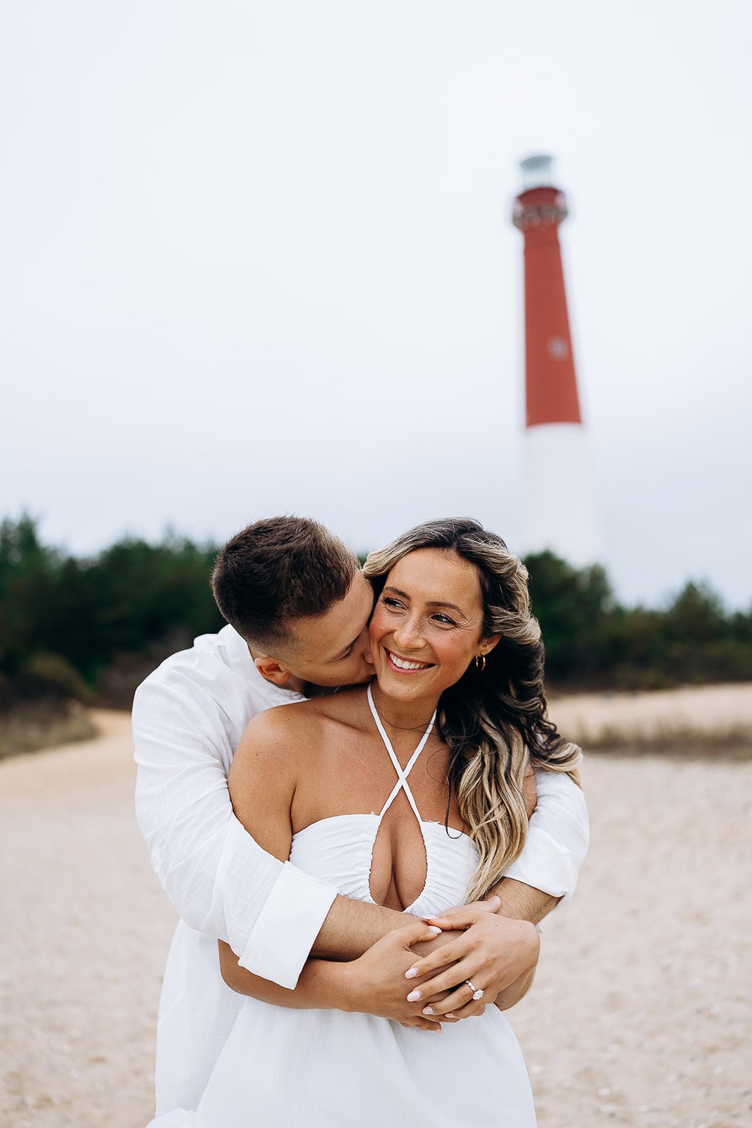 Couple in white attire embracing on sandy path with Barnegat Light lighthouse blurred in the background.