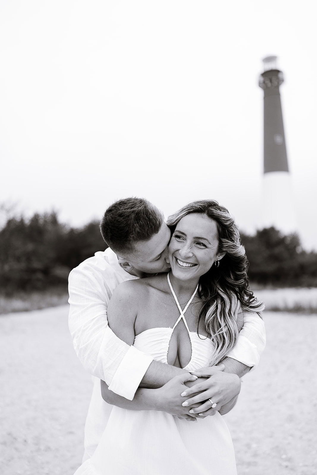 Couple embracing on sandy beach with Barnegat Light lighthouse in the background, woman smiling as man kisses her cheek.