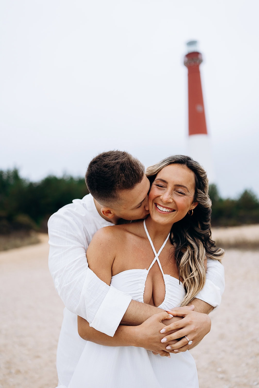 Couple embracing on a sandy path with the red and white Barnegat Light lighthouse in the background, the man gently kissing the woman's cheek.