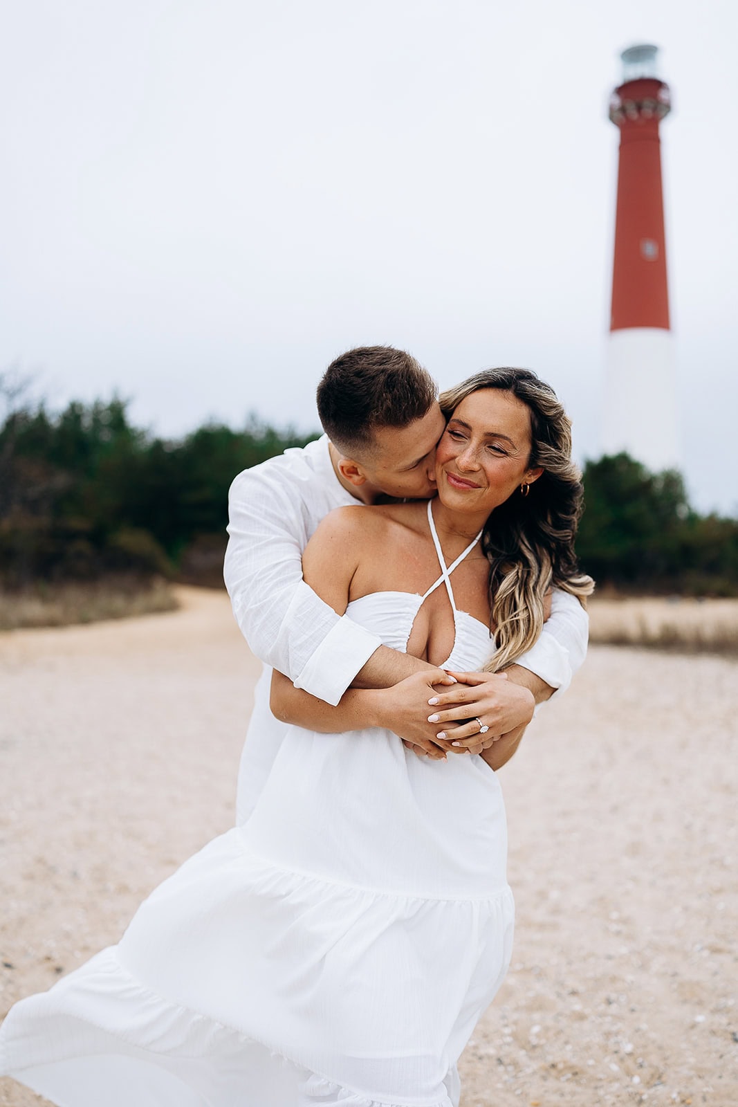 Couple in white clothing embracing on a sandy path with Barnegat Light lighthouse in the background under a cloudy sky.